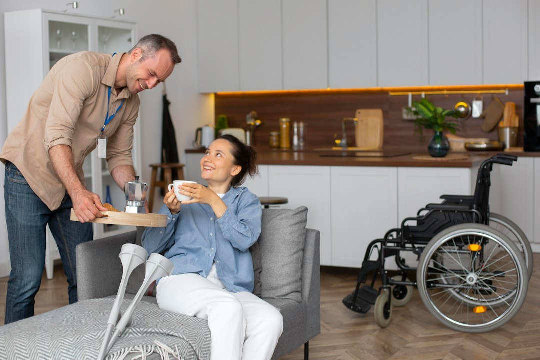A support worker brings tea to a smiling woman on a sofa, with her crutches and wheelchair nearby
