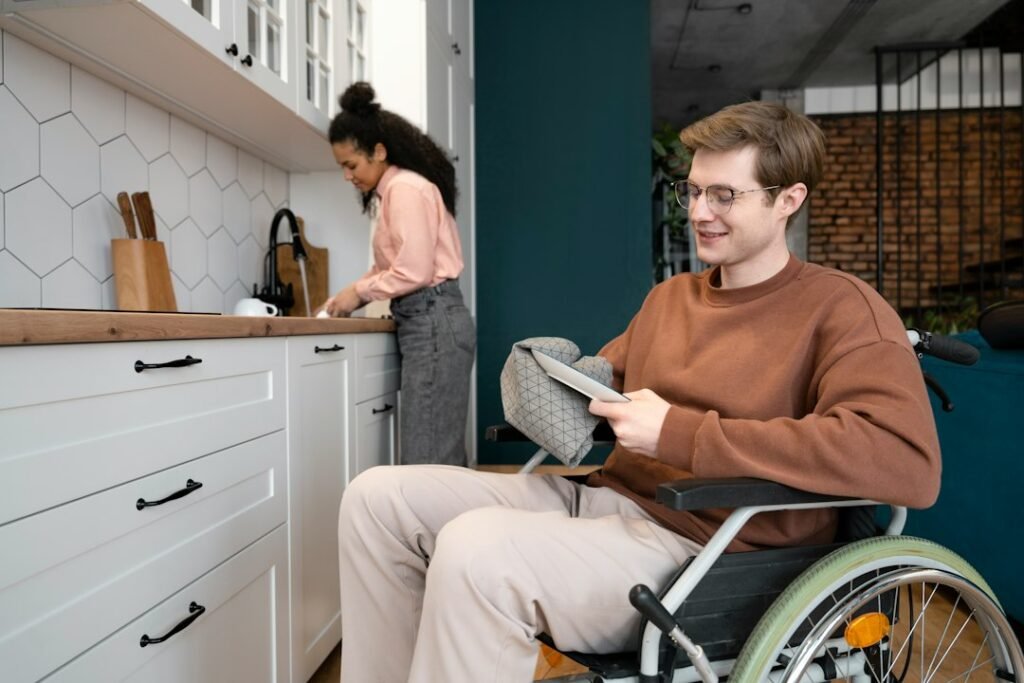 A young man in a wheelchair smiles at a tablet in his kitchen while a woman cooks behind him
