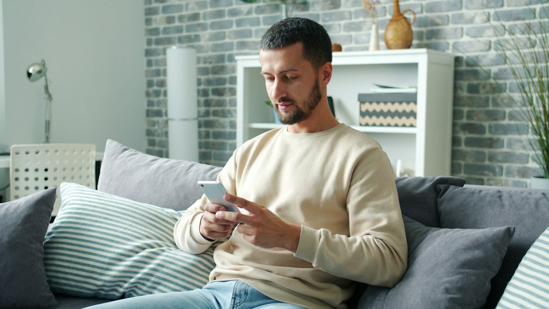 A man sitting on a sofa at home, smiling at his phone